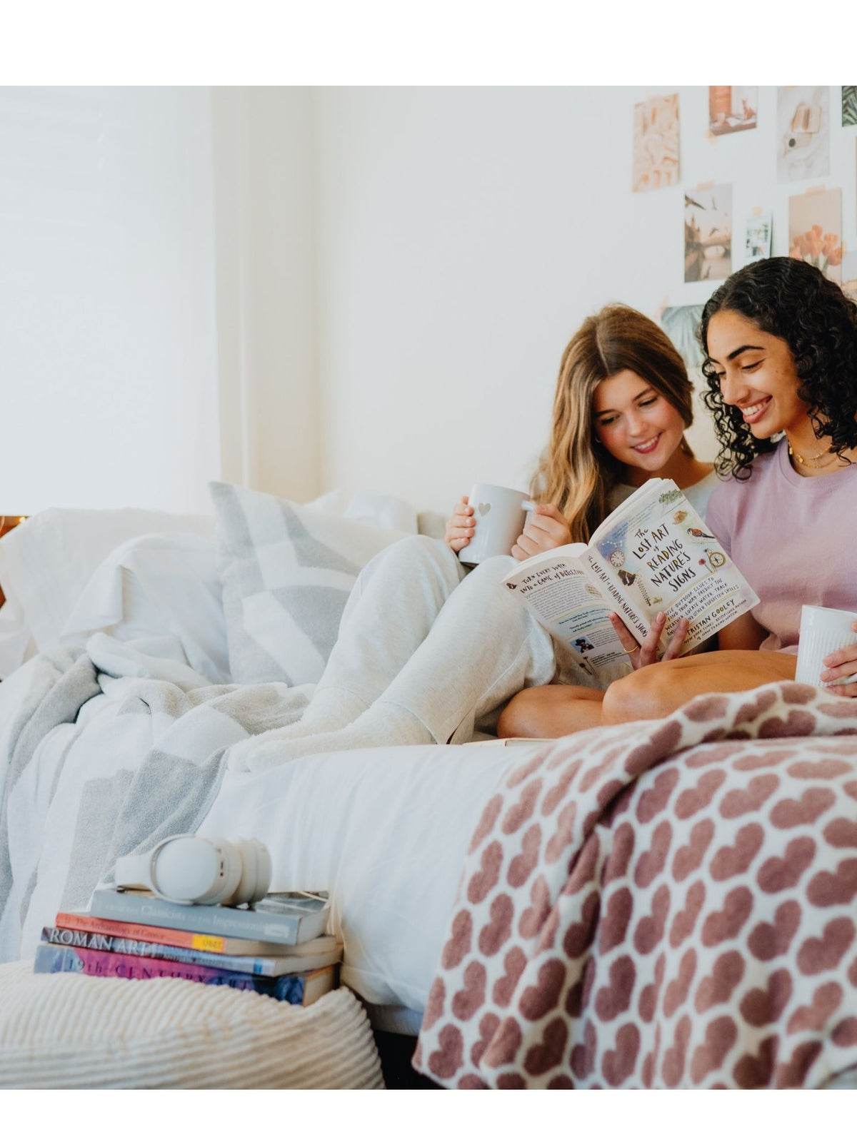 Two women sitting on a bed, one reading a book and the other looking at her.