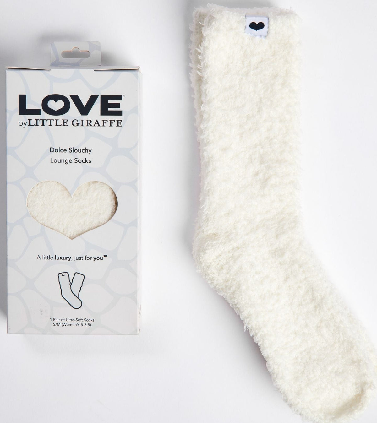 White fluffy lounge sock next to its packaging with 'LOVE by Little Giraffe' branding on a light gray background.