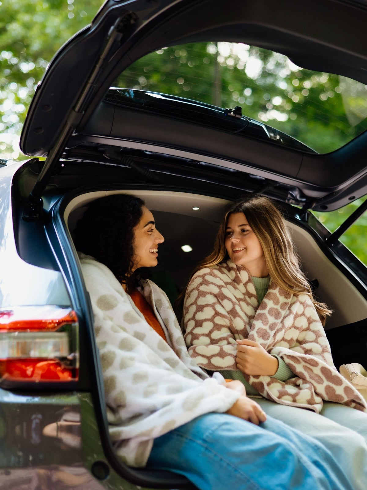 Two women sitting in the back of a car with open trunk, wrapped in a blanket.