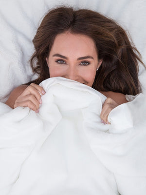 A person holding a beige Luxe™ brand throw over their shoulder, standing against a dark background.
